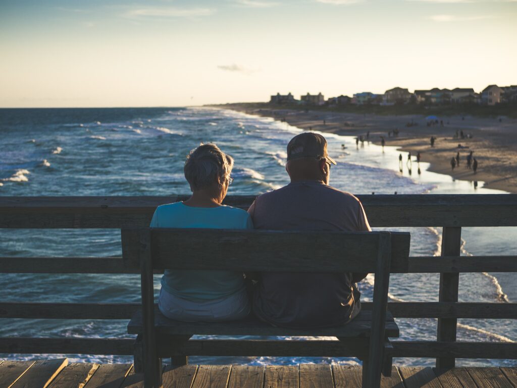 an elderly couple sitting at a bench, watching the water